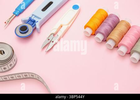 Spools of colorful thread, scissors, rotary cutter and measuring tape neatly arranged on soft pink background. Tools for sewing top view Stock Photo