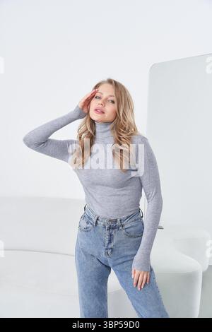 Young woman with long blonde hair wearing a fitted gray turtleneck and high-waisted blue jeans poses confidently, hand on face, in a minimalistic Stock Photo