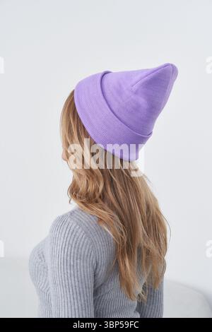 A woman with long blonde hair wears a light purple beanie and a gray ribbed sweater. The image is captured from behind, showcasing the texture of the Stock Photo