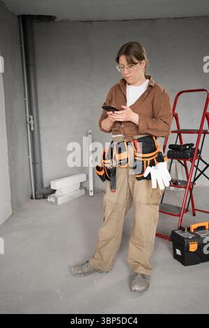 Woman repairman in tool belt in empty apartment making repair works texting and chatting online using smartphone. Renovation, technology and Stock Photo