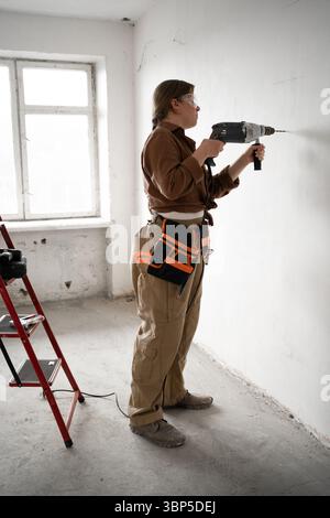 female repairwoman with tool belt drilling wall with hammer drill working on construction site or home renovation Stock Photo