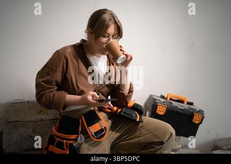 Professional repairwoman in tool belt using smart phone and drinking coffee while resting after work in home Stock Photo
