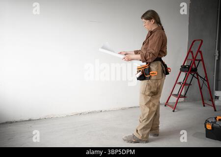 Repairwoman wearing tool belt holding paper blueprint while standing on construction site or inside home under renovation. Concept of DIY repair Stock Photo