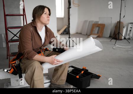 Repairwoman in workwear blueprint sitting at home improvement construction site reading housing development project. Stock Photo