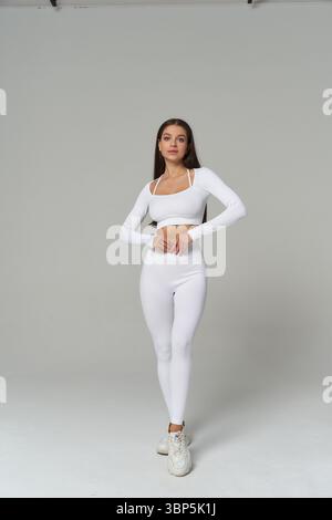 Young woman in a white long-sleeve crop top and leggings, standing confidently while looking forward. She wears white sneakers and poses against a Stock Photo