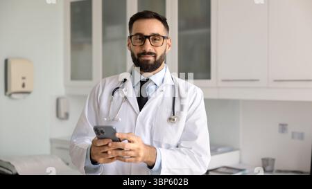 Positive young Hispanic physician man in lab coat holding smartphone Stock Photo