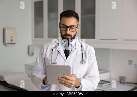 Serious busy Latin doc in stylish eyeglasses typing on tablet Stock Photo