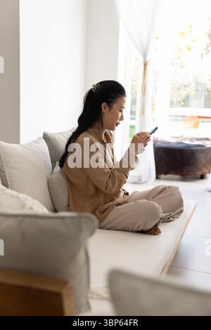 Engrossed Asian woman sits on sofa with smartphone Stock Photo