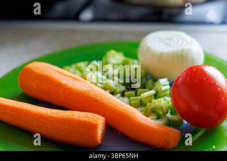 Fresh vegetables are displayed on a green plate, including two peeled carrots, sliced ​​celery stalks, half an onion, and one tomato. Stock Photo