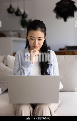 Engrossed Vietnamese woman working on laptop seated on couch Stock Photo