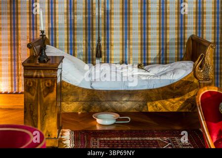 A historic bedroom in Arenenberg Castle in Salenstein, Thurgau, Switzerland, features a bed with ornate carvings, a bedside table with a candlestick and a chamber pot on the wooden floor. The room, once occupied by Hortense de Beauharnais and her son Napoleon III, provides an insight into 19th century home decor Stock Photo