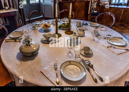 A fully laid, round dining table stands at Arenenberg Castle in Salenstein, Thurgau, Switzerland. Limoges porcelain bearing a crowned "N" monogram, gold candelabra, tureens, a gravy boat and a fruit bowl are arranged for a festive meal. Dark wooden furniture surrounds the room. Stock Photo