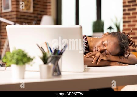 Overworked black businesswoman falls asleep at desk in home office after completing numerous tasks on laptop. Exhausted remote employee naps while seated, recovering from long hours of intense work. Stock Photo