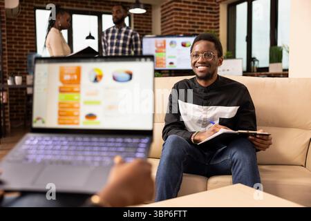 Two business professionals meet in office to share ideas and collaborate on project task. African american sales manager with laptop presents commercial offer to startup company client seated on sofa. Stock Photo