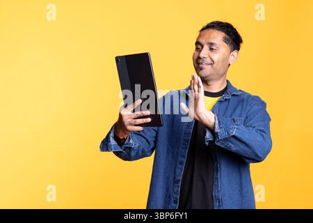 Indian person attending an online videoconference on his tablet in studio, smiling and chatting with his friends against orange background. Young adult waving hand with a positive mood. Stock Photo