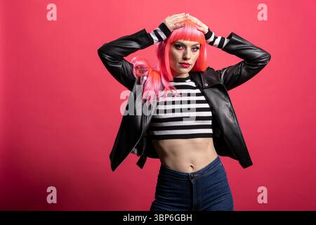 Trendy model with pink hair and bold jacket embraces confident rocker style against isolated background. Pretty woman exudes funky attitude and stands gracefully, expressing punk vibe and chic flair. Stock Photo