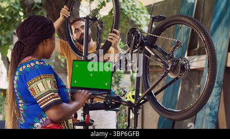 Sports-loving couple in yard repairs bicycle with laptop that is displaying isolated chromakey template. Girlfriend carrying pc with blank copyspace screen while boyfriend safely secures bike wheel. Stock Photo