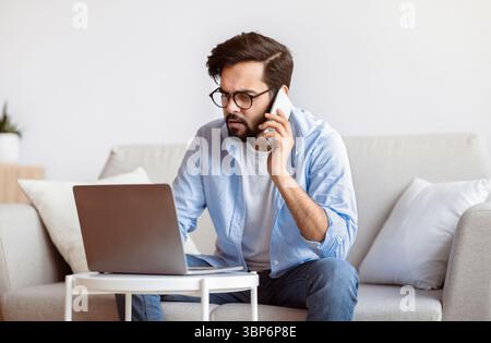 Busy Eastern Freelancer Guy Talking On Phone And Using Laptop At Home Stock Photo