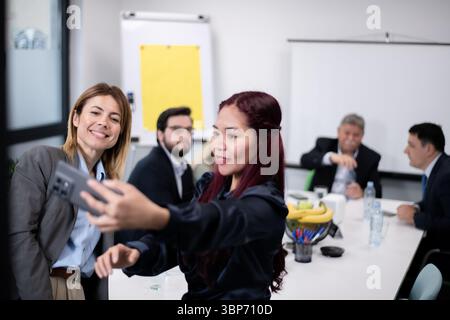 A group of business professionals interacting in a bright office, focusing on two colleagues taking an engaging selfie and others discussing around a Stock Photo