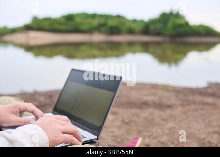 Unknown person sitting outdoors on the bank of a stream, working on his laptop. Concept: digital nomad working remotely while traveling, enjoying a li Stock Photo