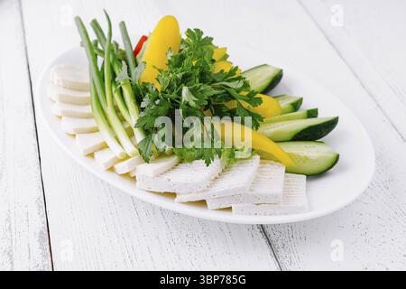 Food, White plate is sitting on a white wooden table with sliced cheese, cucumber, peppers, and green onions arranged on top Stock Photo