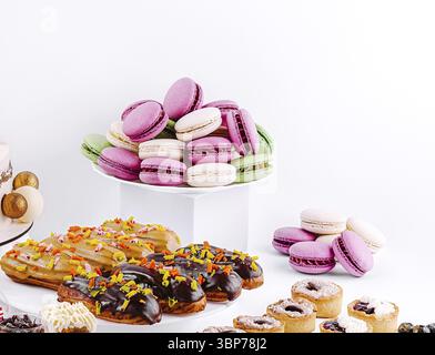 Food, A variety of macarons in pastel colors are stacked on a white pedestal. Nearby, assorted pastries including donuts and eclairs are arranged arti Stock Photo