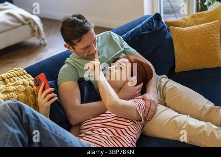Couple in love lying on sofa with smartphone and hugging while bonding at home. Young family woman and man resting, doing nothing together. Staying co Stock Photo