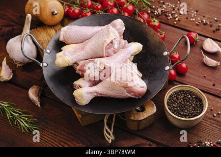 Food, A selection of raw chicken drumsticks is displayed in a black skillet alongside cherry tomatoes and spices on a wooden table, showcasing an invi Stock Photo