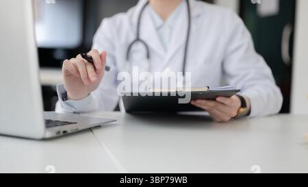 Close up of doctor with stethoscope holding fountain pen while sitting in front of laptop at white table in diagnosis room, Hospital or Clinic office. Stock Photo