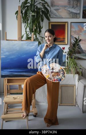 Satisfied artist woman smiling looking at camera holding colour palette stands near easel with canvas picture in art studio. Joyful female painter wit Stock Photo