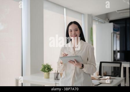 Female office worker is holding a stylus writing on tablet while standing in front of working table. Artist or Designer, Modern company job. Stock Photo