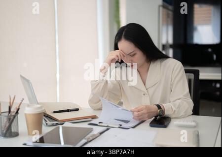 Businesswoman or female office worker reading documents with a hand touching her temple while sitting at working table. Corporate Job, Modern company. Stock Photo