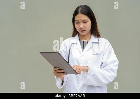 Young Asian woman doctor wearing white lab coat and stethoscope against plain background Stock Photo