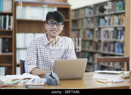Young Asian male university student doing research inside a library alone. Young man sitting and working on a project. Focused man reading and writing Stock Photo