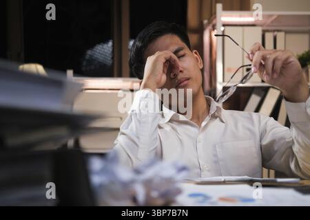 Exhausted young man with laptop in office working late sitting on desk in office overtime at night. Frustrated young businessman massaging his nose an Stock Photo