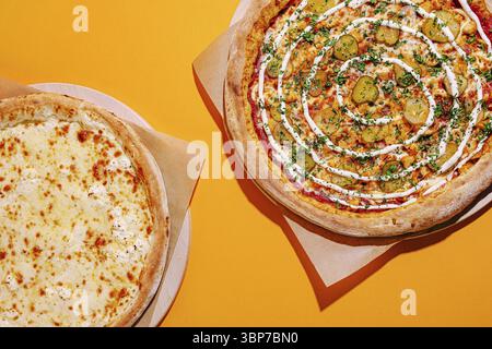 Food, Two pizzas are presented on light brown paper, one with cheese and the other topped with pickles and herbs. The background is a bright orange, e Stock Photo