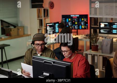 Two young adult men wearing headsets sitting at desk discussing script while working on computer in scam call center, background showing monitors Stock Photo