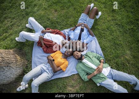 Group of thoughtful relaxed young African American friends lying together on green grass outdoors. Top view of four black students resting on plaid in Stock Photo