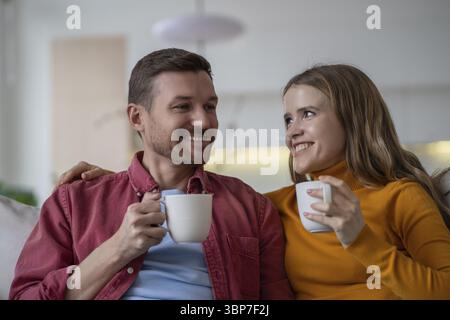 Happy couple man and woman drinking tea coffee sitting on sofa at home, enjoying talking tender hugging. Loving spouses wife husband bonding having ni Stock Photo