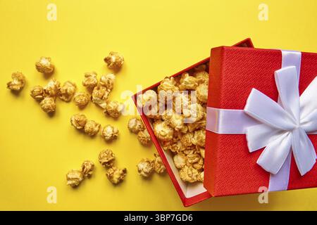 Red gift box with white bow full of tasty caramel popcorn on yellow background Stock Photo