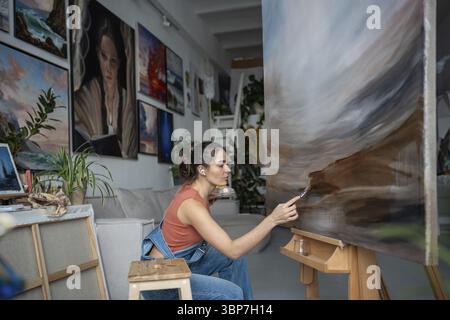 Talented serious woman artist working on abstract painting with careful gentle strokes of small brush. Girl wear earphones creating dark impressionist Stock Photo