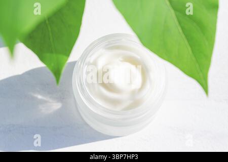 Moisturizer cream jar in frosted glass bottle and green leaves on white background. Close up of skin and body care beauty products texture Stock Photo