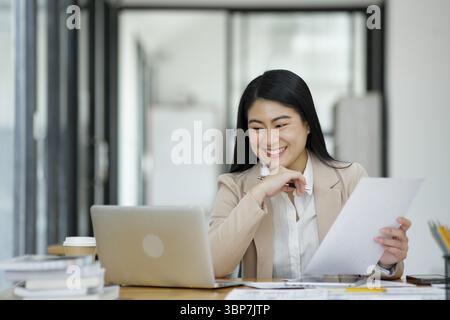 A focused businesswoman working on a laptop while holding a notebook in a bright office environment Stock Photo