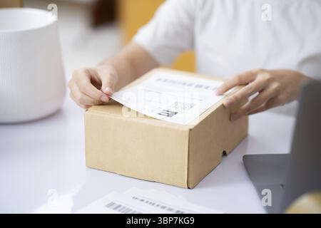 Closeup Male entrepreneur startup small business owner packing package preparing delivery parcel and attaching data label on carboard box on desk. Owe Stock Photo