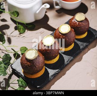 Food, A set of four round desserts is arranged on a black slate plate. Their rich chocolate coating glimmers under soft lighting, with a teapot and cu Stock Photo
