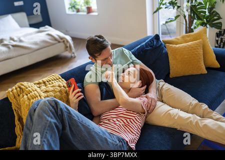 Happy couple spend time together at home. Girl touches boyfriend nose cutely. Lover fooling around on the couch. Girl lies on lap of guy and looks int Stock Photo