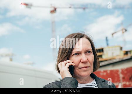 Portrait of elegant brunette businesswoman using mobile phone on construction site, selective focus Stock Photo