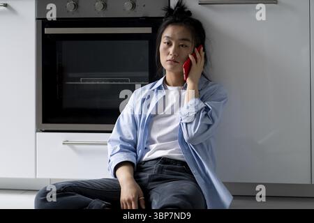 Depressed young Asian woman sitting on floor at home constantly calling ex-boyfriend after breakup, feeling lonely, talking with close friend over pho Stock Photo