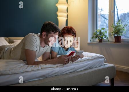Couple looking phone while lying on bed. Happy boyfriend and girlfriend watching funny video on mobile and smiling, checking social media. Man showing Stock Photo