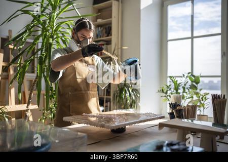 Serious female artist works with epoxy resin, creating picture in technique of liquid art. Professional woman wielding mixing stick, carefully pouring Stock Photo